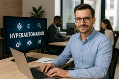 A smiling professional sits at a desk with a laptop and monitor displaying “HYPERAUTOMATION,” with colleagues working in the background of a bright office environment.